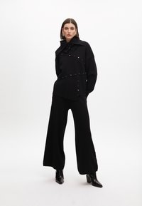 Top negro de gran tamaño con cuello alto, tachuelas doradas a lo largo de las costuras y pantalones anchos negros a juego, combinados con botas de tacón negras.