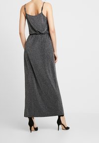 Robe maxi en tissu noir et argent parsemé, avec de fines bretelles spaghetti, une taille élastique et une fente sur le côté, associée à des escarpins noirs.