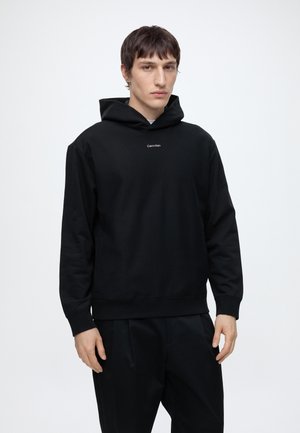 Man draagt een zwarte hoodie met de tekst "Calvin Klein" op de borst, staat frontaal tegen een effen lichte achtergrond, handen ontspannen langs het lichaam.