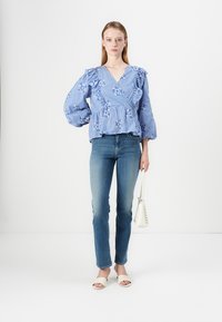 Blouse en vichy bleu à manches volantes et broderies florales, associée à un jean ajusté et des sandales à talons blancs. Tenant un petit sac à main blanc.