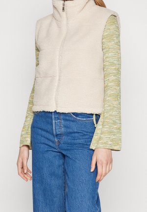 Femme portant une veste polaire crème zippée sur un haut vert à motifs à manches longues et un jean bleu taille haute sur un fond uni.