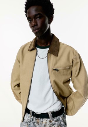 Young man with short curly hair wearing a beige jacket over a white t-shirt and patterned pants, standing with hands in pockets against white background.