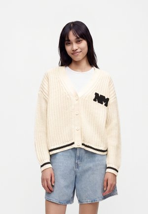 Jeune femme aux cheveux foncés portant un cardigan crème en maille boutonné avec des rayures noires et un short en jean bleu clair, souriant légèrement.