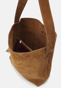 Sac fourre-tout en daim marron avec une texture douce, un dessus ouvert et une petite poche intérieure zippée. Dispose de bretelles épaisses solides et d'un design minimaliste.