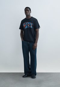 Hombre de pie contra una pared lisa, vistiendo una camiseta negra con el gráfico "Billionaire Boys Club", jeans oscuros y sueltos, y zapatos negros.