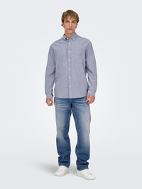 Jonge man staat, gekleed in een lichtblauw overhemd met knopen, vervaagde blauwe spijkerbroek en witte sneakers, tegen een effen lichte achtergrond.