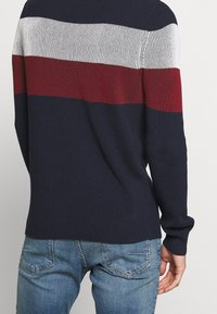 Marinblå ribbad tröja med horisontella ränder i vitt och vinrött. Har långa ärmar och rund halsringning. Bärs med blå jeans.