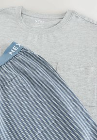 Tricou gri cu mânecă scurtă și buzunar, purtat cu pantaloni scurți albaștri cu dungi și talie solidă, inscripționată "NEXT."
