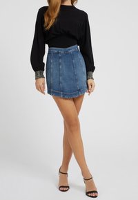 Femme portant un haut noir à manches longues avec des poignets ornés, une mini-jupe en jean bleue et des talons ouverts noirs, debout sur un fond blanc.