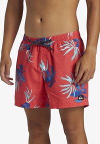Red swim shorts with blue and light blue tropical leaf patterns, an elastic waistband with a drawstring, and a small logo patch.