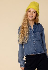 Leren button-up overhemd, lichtblauw met een zak, cropped fit, gecombineerd met zwarte broek. Geaccentueerd met een felgele gebreide beanie.
