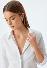 Camicia bianca di cotone con colletto a punta, sbottonata al collo. Tessuto morbido; il modello indossa orecchini a cerchio in argento.