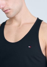 Svart tanktop med rund hals og en liten brodert logo i rødt, hvitt og blått på venstre bryst. Glatt bomullstekstur.