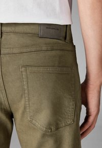 Oliivroheline denimipüksid, millel on tagumine tasku ja nahasilt, millel on kiri "DENIM SUPPLY Co." koos peente tekstuuri ja õmbluse detailidega.