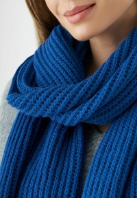Femme portant une épaisse écharpe en tricot bleu texturé enroulée autour du cou sur un pull gris, montrant le bas du visage et le menton.