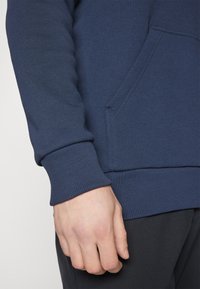 Sweat à capuche bleu marine en matériau doux, avec une poche avant, des poignets côtelés et une coupe décontractée. Tissu texturé avec des détails de coutures minimes.