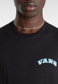 Černé tričko s krátkým rukávem, které má na levé části hrudi světle modrý klenutý nápis "VANS". Hladká tkanina s klasickým kulatým výstřihem.