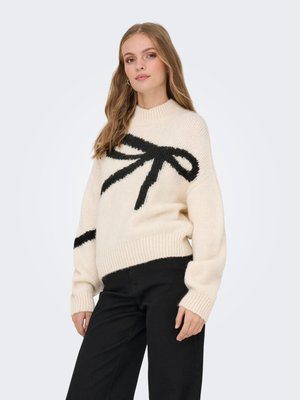 Femme portant un pull crème avec un grand motif de nœud noir et un pantalon noir, debout devant un fond clair uni.
