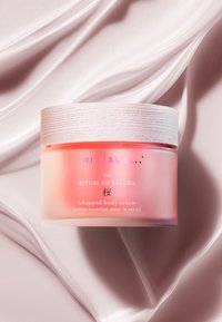 Vispad kroppskräm i en frostad rosa burk med ett texturerat trämlock, med vit och silver accenttext. Slät rosa bakgrund.