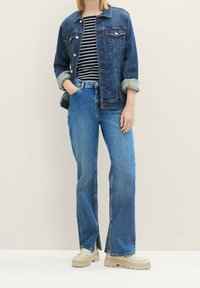 Denimjacke, hellblau, mit Knöpfen und zwei Brusttaschen. Gestreiftes Schwarz-Weiß-Oberteil darunter. Hellblaue Schlagjeans, beigefarbene Schuhe.