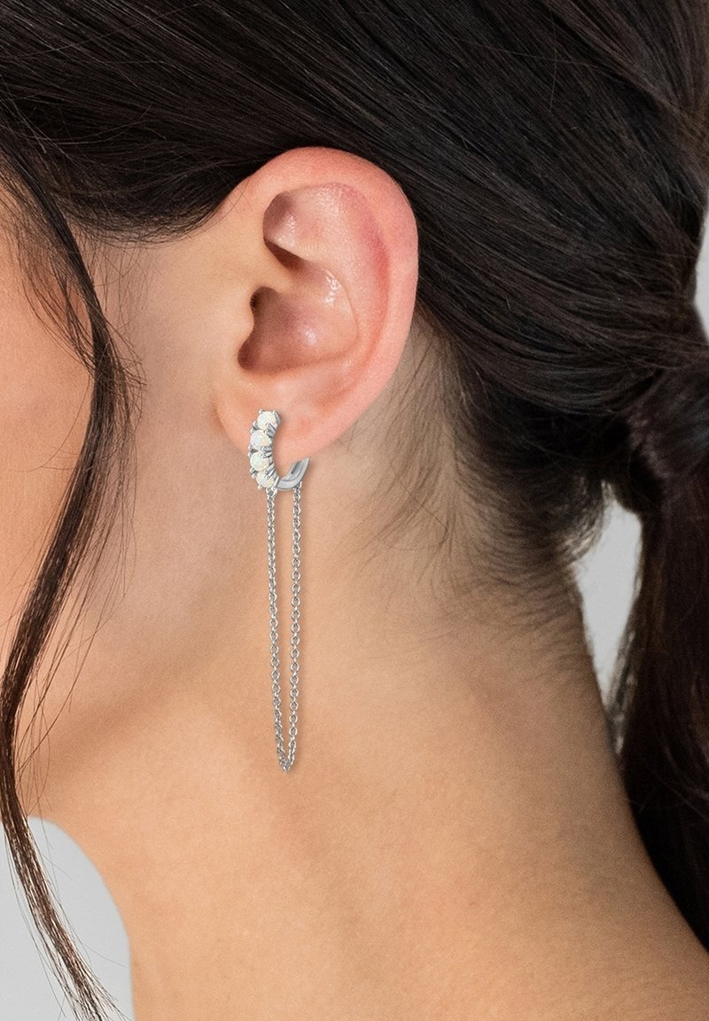 Sudraba riņķveida auskari ar pieciem baltiem opāla līdzīgiem akmeņiem un garu, smalku ķēdīti, kas karājas zemāk. Virsma ir teksturēta un pulēta.