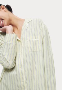 Camisa de botones de rayas amarillo claro con finas líneas marinas, de manga larga, con collar y un bolsillo en el pecho. Textura de tela suave.