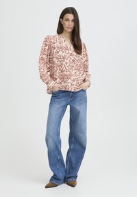 Une femme se tient avec les mains dans les poches, portant un blouse à manches longues à motifs floraux, un jean bleu à larges jambes et des chaussures brunes à bouts pointus, sur fond blanc.