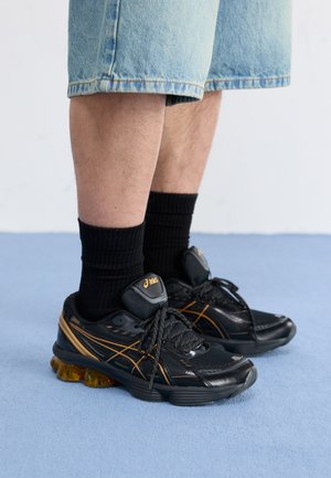 Juodos sportinės kedės su oranžinėmis detalėmis, tekstūruotas viršus ir paminkštintas padas. Nešiojamos su juodomis dryžuotomis kojinėmis ir šviesiai mėlyniais džinsiniais šortais.