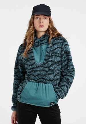 Faux-Fur-Pullover in Teal mit schwarzen Wellenmustern, verfügt über einen halben Reißverschluss-Kragen und eine einfarbige tealfarbene Kängurutasche. Eine schwarze Mütze vervollständigt den Look.