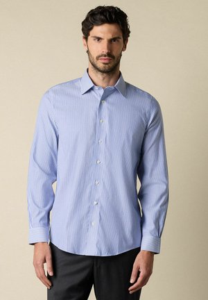 Man met donker haar en baard, gekleed in een lichtblauw-wit gestreept overhemd met lange mouwen en donkere broek, voor een neutrale achtergrond.