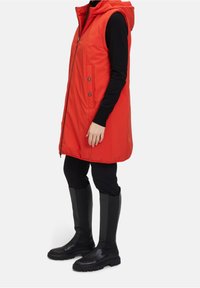 Veste rembourrée rouge avec capuche, fermeture éclair à l'avant et boutons-pression, poches latérales, assortie à un haut noir ajusté et des bottes noires montantes.