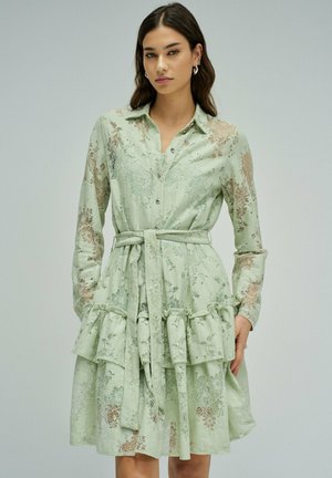 Mujer con un vestido de encaje verde claro con mangas largas, cintura ceñida con cinturón y capas de falda con volantes sobre un fondo liso.