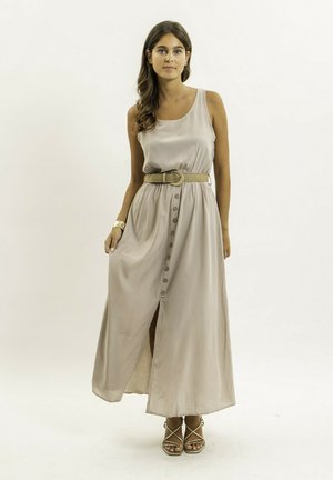 Femme aux longs cheveux foncés portant une robe maxi beige sans manches avec des boutons et une large ceinture, debout avec une expression neutre sur un fond clair.