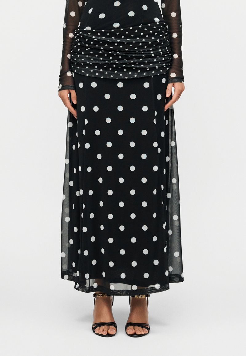 Μαύρο φόρεμα με λευκά πουά, ραμμένο με πιέτες στη μέση, διαφανή μανίκια και ροή στη φούστα. Αξεσουάρ περιλαμβάνουν μαύρα σανδάλια με λουράκια και χρυσές λεπτομέρειες.