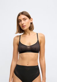 Sort bralette med et geometrisk mønster, gennemsigtig stof og justerbare stropper. Har en logo-elastik. Sammen med højlavede sorte gennemsigtige trusser.