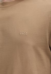 Chemise marron clair avec un petit texte brodé centré « BOSS » sur la partie supérieure de la poitrine, gros plan sur la texture du tissu et les coutures.