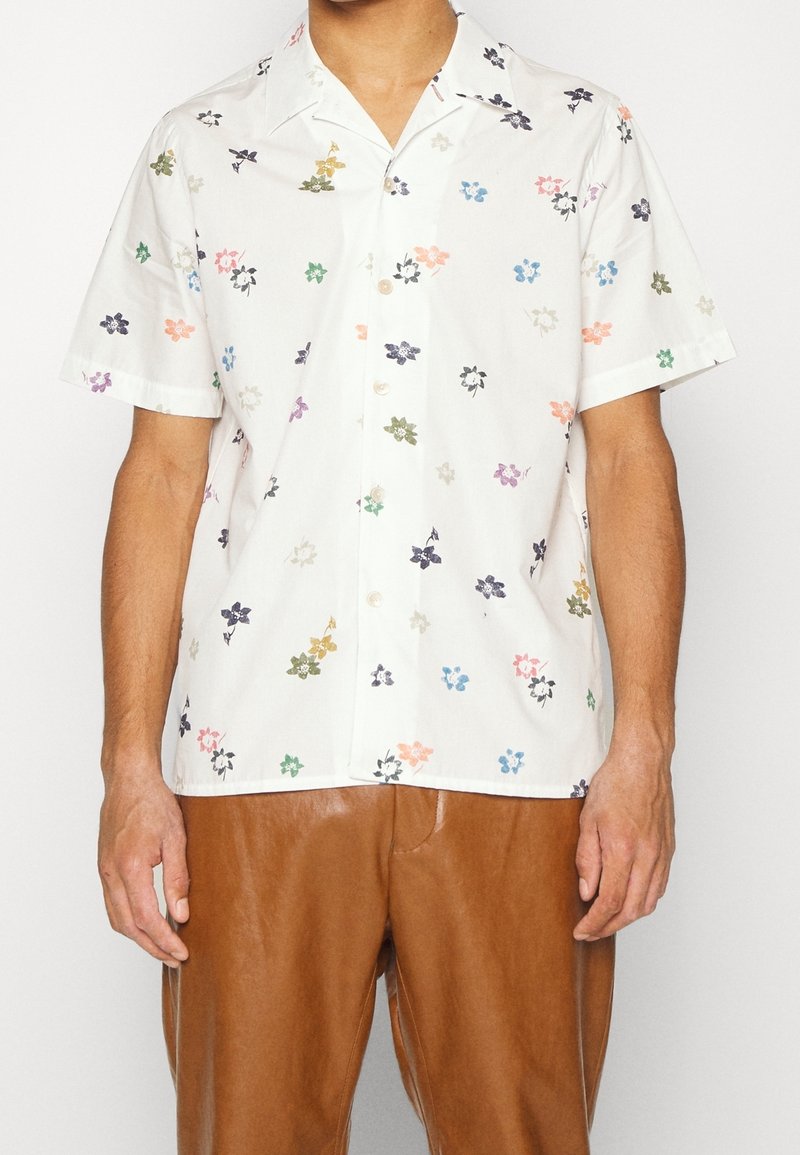 Homme portant une chemise blanche à manches courtes avec un petit imprimé floral multicolore et un pantalon en cuir marron, debout devant un fond uni.
