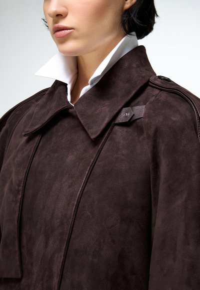 Közeli kép egy személyről, aki sötétbarna velúr kabátot visel széles gallérral, fehér, felálló galléros ing fölött.