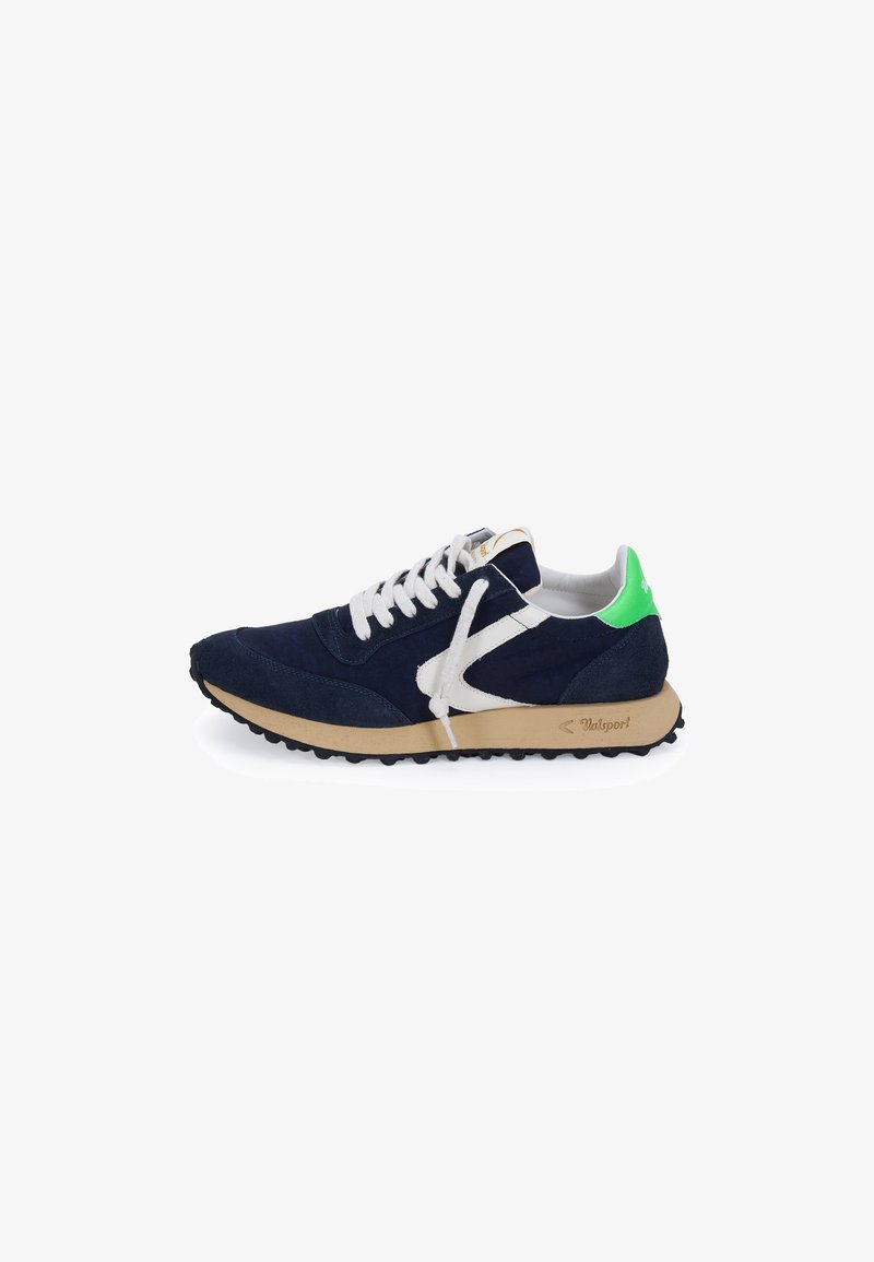Zapatilla deportiva azul marino con cordones blancos, suela beige, logo lateral blanco y lengüeta de talón verde brillante, mostrada de perfil sobre fondo blanco.