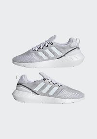 Szürke kötött sportcipők, három fehér csíkkal, fekete részletekkel, fehér talppal és texturált felsőrésszel. Kerek fűzőkkel és húzófüllel rendelkeznek.