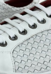 Las zapatillas blancas de cuero presentan un diseño tejido, ojales plateados y cordones blancos. El interior tiene un forro rojo en contraste.