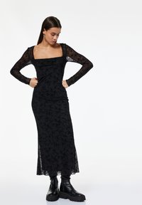 Frau in einem langen schwarzen Blumenspitzenkleid mit durchsichtigen langen Ärmeln, die schwarze klobige Stiefel trägt, steht mit den Händen in den Hüften und blickt nach unten.