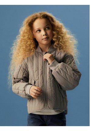 Jauna mergaitė su ilgais garbanotais šviesiais plaukais, dėvinti šviesiai pilką siuvinėtą striukę, laiko užtrauktuką, stovėdama prie mėlyno fono.