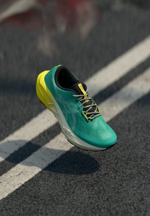 Pantof de alergare verde și galben în aer deasupra unui drum asfaltat cu marcaje albe, evidențiind mișcarea și detaliile designului sportiv.