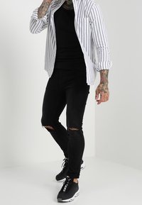 Gestreepte overhemd in wit en marineblauw, zwart T-shirt met ronde hals, gescheurde zwarte skinny jeans en zwarte sneakers met witte zolen.