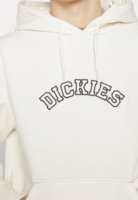 Sudadera crema con un bolsillo canguro en la parte frontal; presenta un logo bordado en negro de "DICKIES" en forma de arco. Textura suave y capucha con cordones.