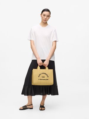 Nainen seisoo valkoisessa t-paidassa, mustassa laskostetussa midihameessa ja mustissa sandaaleissa, pitäen keltaista Karl Lagerfeldin kangaskassia kahvoista kiinni.