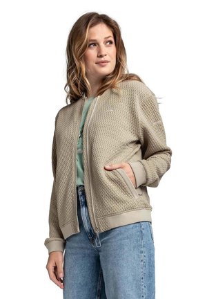 Hellbeige Bomberjacke mit strukturiertem Obermaterial, Reißverschluss, gerippten Bündchen und Saum sowie seitlichen Taschen, kombiniert mit einem grünen Oberteil und Jeans.