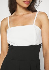 Femme portant un haut blanc sans manches avec des fines bretelles et un pantalon noir taille haute avec un détail plissé à la taille.