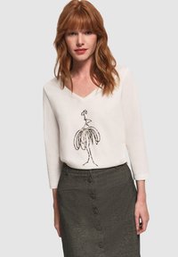 Donna con capelli rossi che indossa un top bianco a scollo a V con un design astratto di uccello in nero e una gonna grigia a vita alta con bottoni.